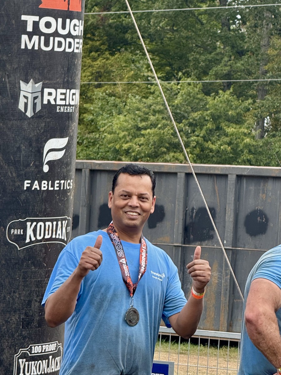 Arvind Choudhary smiling with two thumbs up, wearing a Tough Mudder finisher's medal at the finish line.